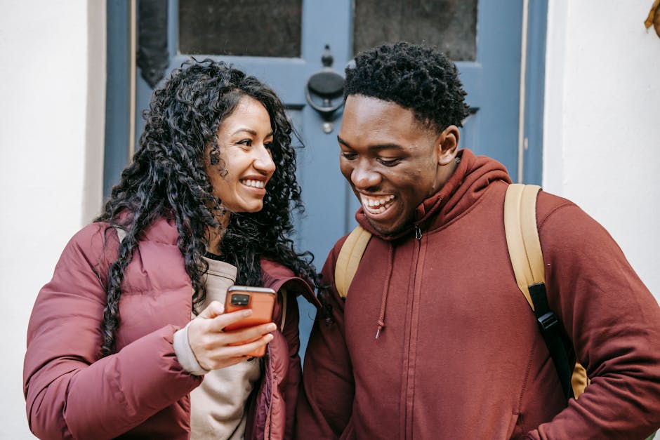 Young happy multiethnic partners talking while browsing internet on cellphone in town in daytime