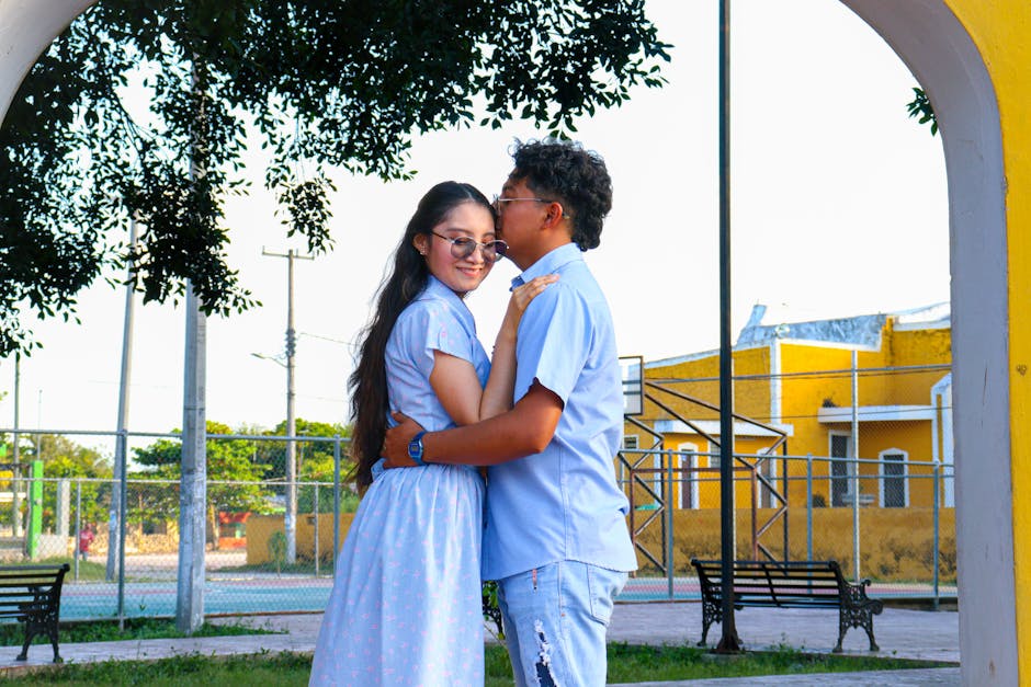 A young couple in casual attire, embracing and smiling under a park archway.