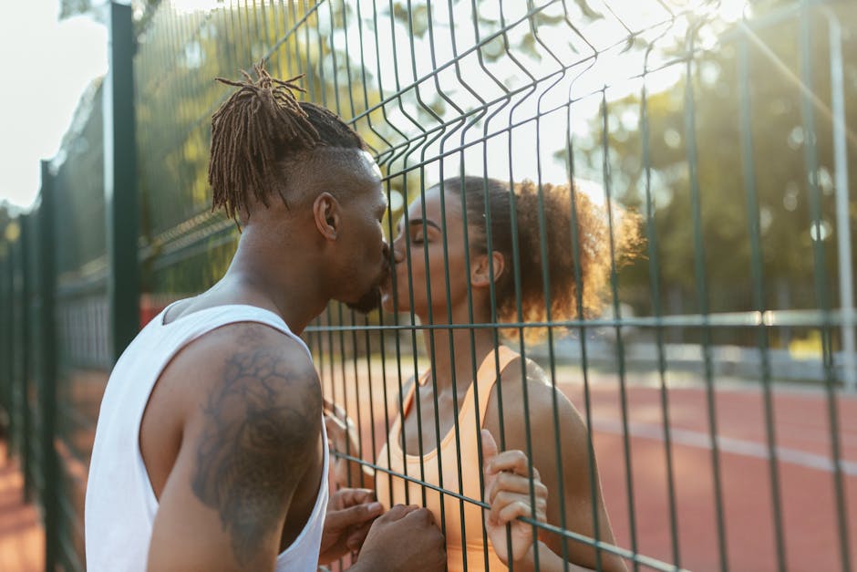 A couple shares a tender kiss through a fence on a sunny day, highlighting love and connection.