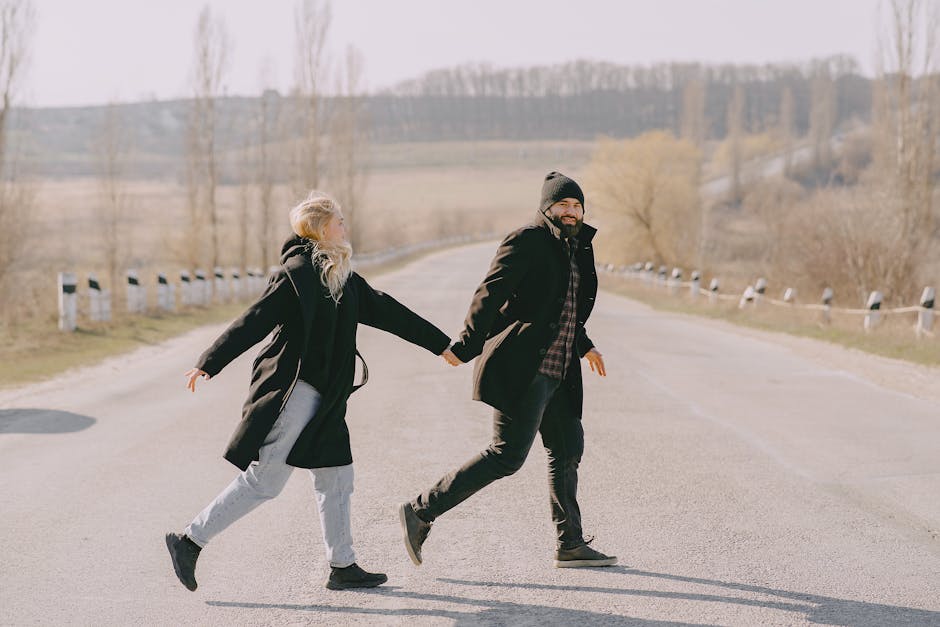 Side view full body of cheerful couple in casual warm outerwear holding hands and crossing road in rural area in spring morning