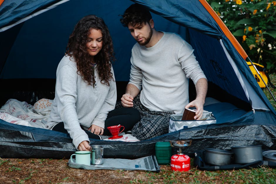 A young couple enjoys coffee inside a tent during a camping trip, surrounded by nature.