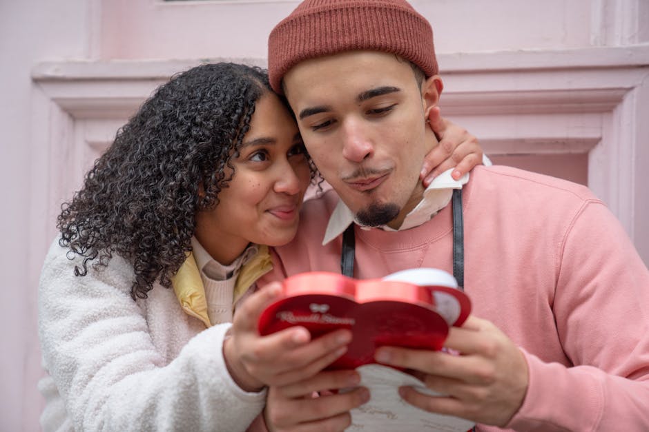 Joyful young ethnic couple embracing and eating yummy candies from heart shaped gift box during anniversary celebration