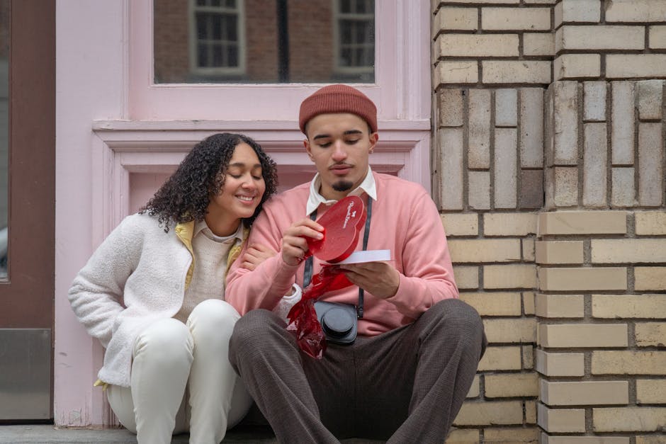 Young ethnic guy in stylish outfit opening heart shaped box with chocolate while sitting on street with happy excited girlfriend on Saint Valentines Day