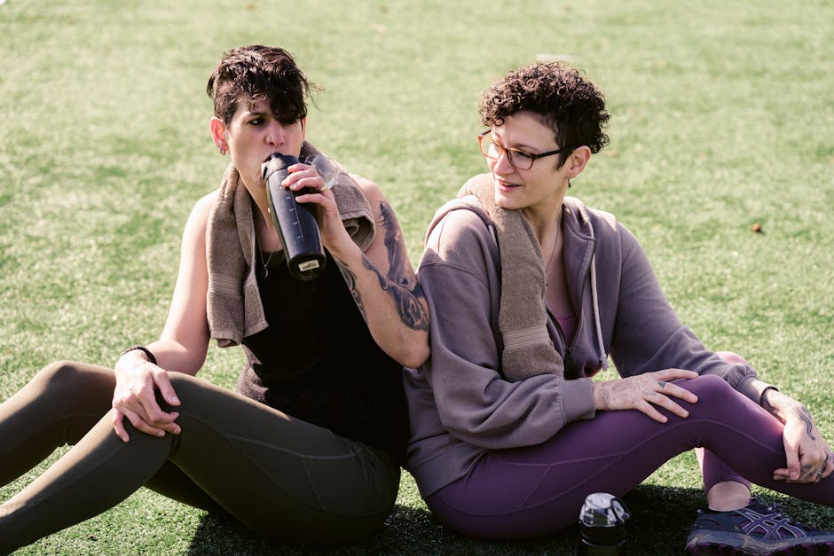 Two women in sportswear relaxing with water bottles in a sunny park.