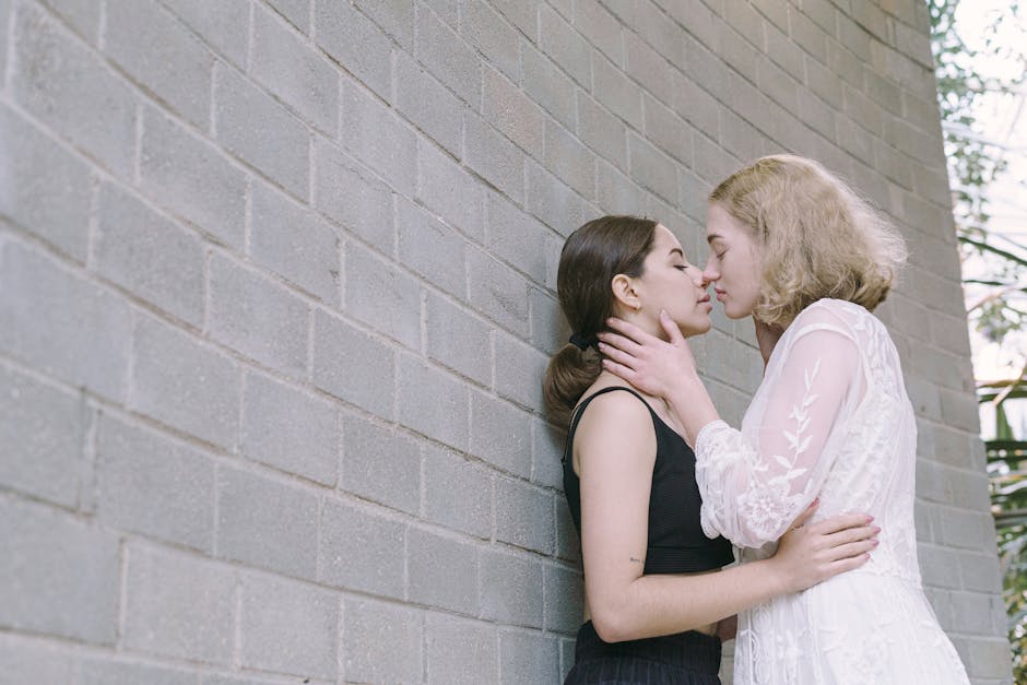 Two young women embracing and sharing a tender moment against a gray brick wall outdoors.