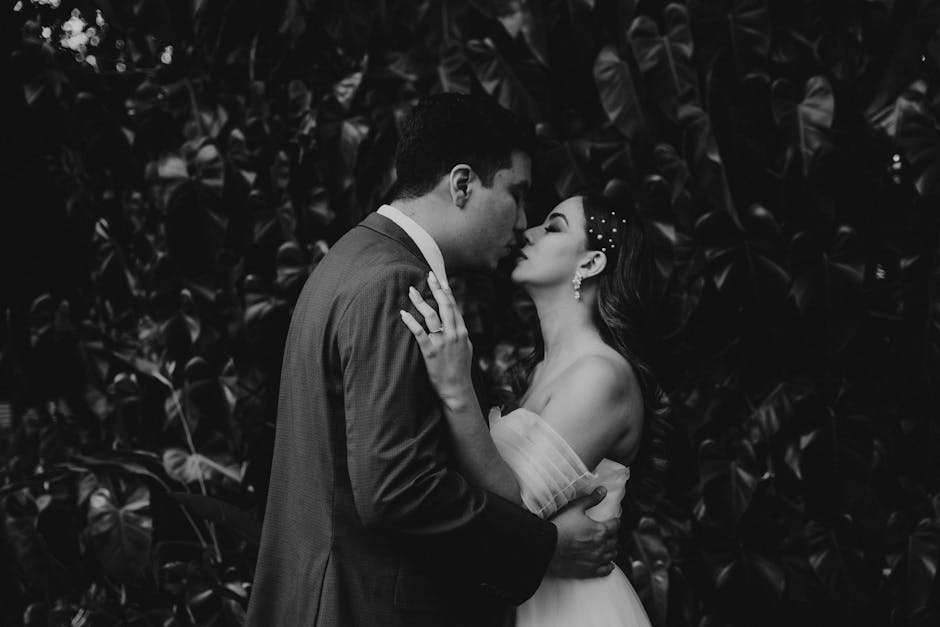 A romantic black and white photograph of newlyweds sharing a tender kiss amidst lush foliage.