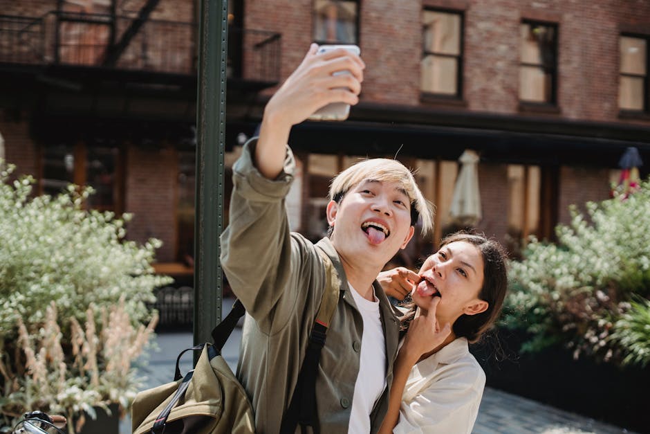 Playful young couple sticking tongues out while taking an outdoor selfie in a sunny urban setting.
