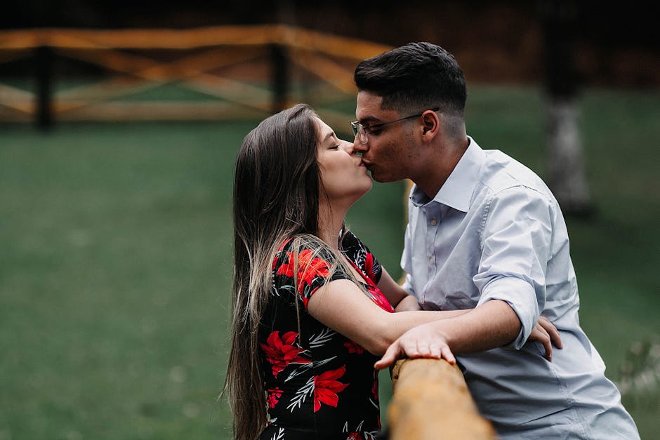 A young couple shares a romantic kiss by a wooden railing in a serene outdoor setting.
