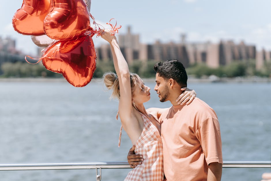 Couple celebrating love by a river, holding heart-shaped balloons on a sunny day.