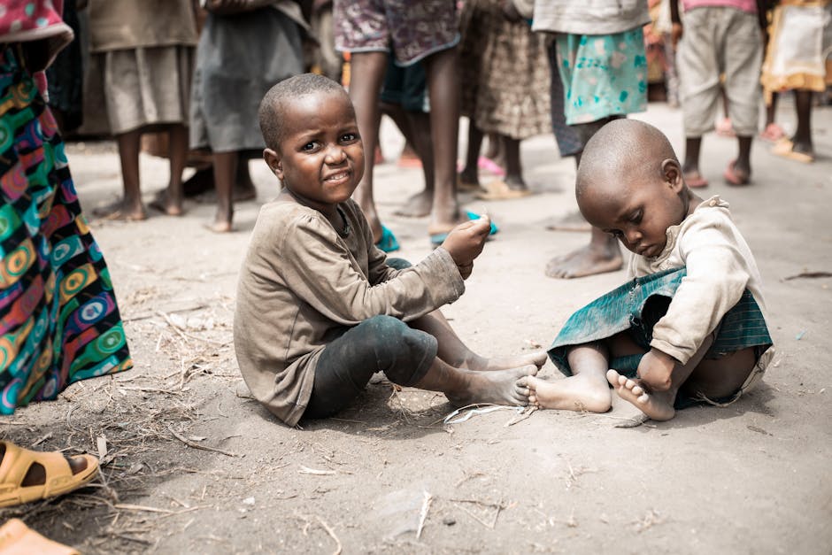 Two young children sitting on the ground in a rural setting, surrounded by people.