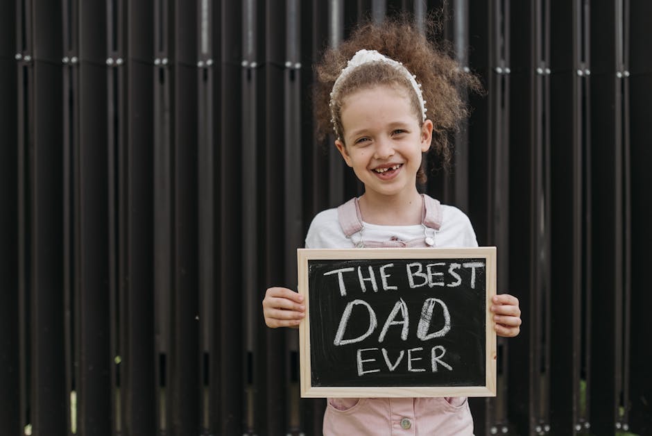 Cheerful young girl holding a blackboard with 'The Best Dad Ever' sign in a playful outdoor setting.