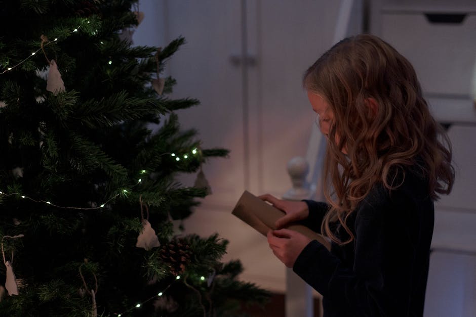 A young girl with wavy hair opens a package beside a decorated Christmas tree indoors.