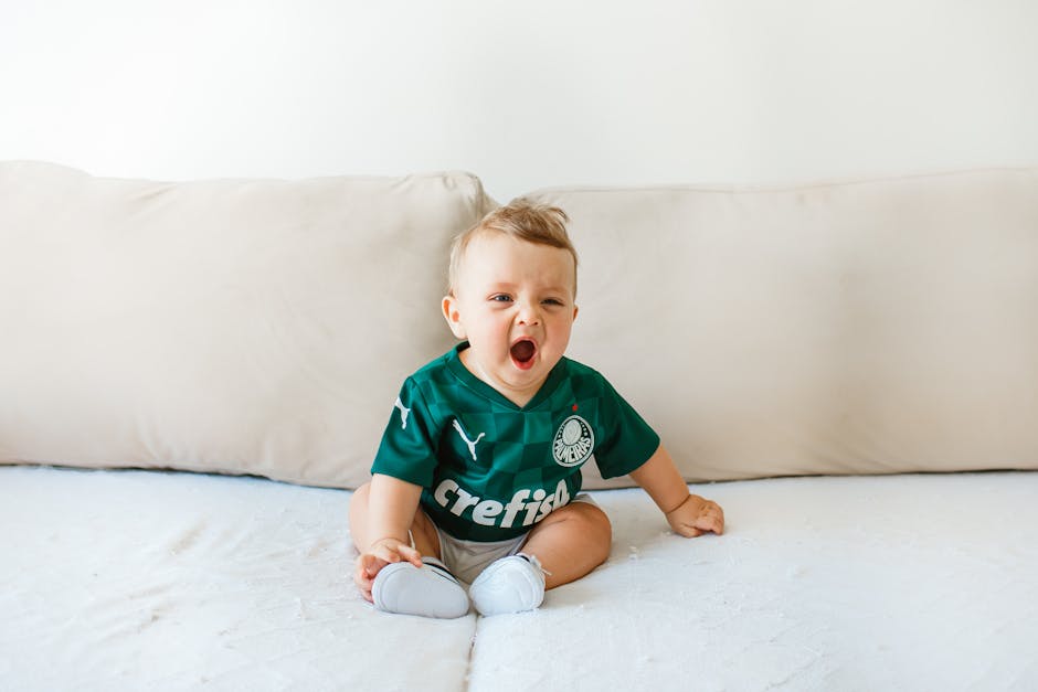 Cute baby wearing a green jersey yawning on a soft beige couch indoors.