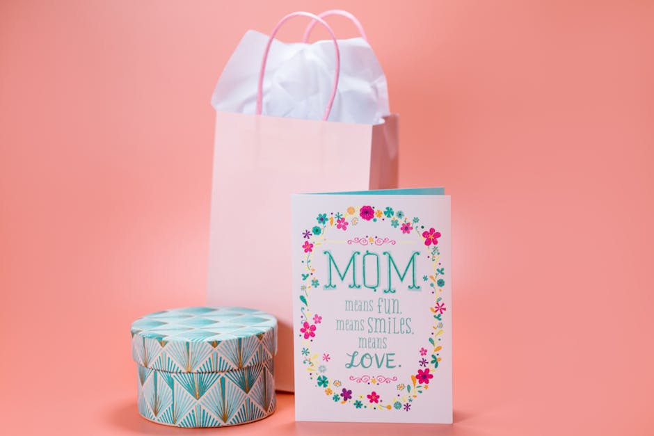 Elegant Mother's Day greeting card with gift bag and decorative box on pastel pink background.