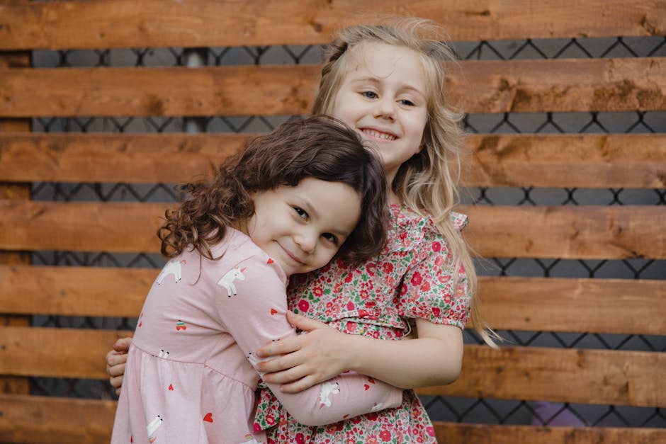 Two young girls smiling and hugging against a wooden fence, wearing floral dresses.