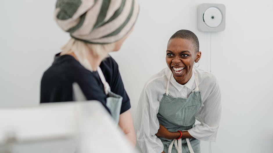 Two women with aprons share a joyful moment, laughing together in a casual indoor setting.
