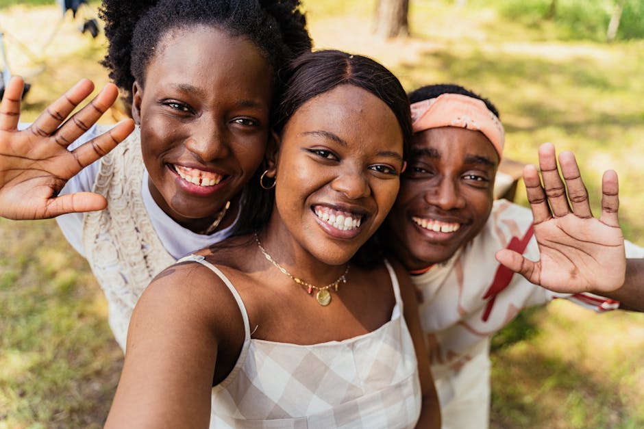 Three young friends capture a joyful outdoor selfie with bright smiles and waving hands.