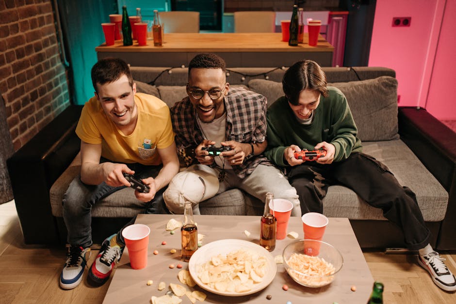 Three men playing video games together on a sofa with snacks and drinks around.