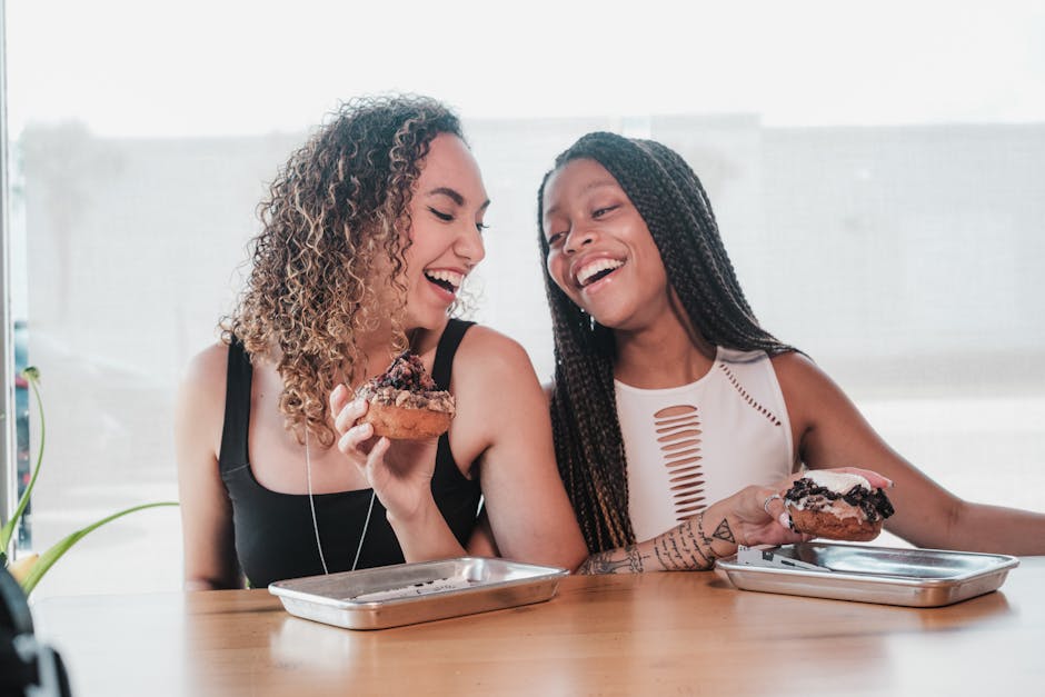 Two smiling women with donuts sharing a joyful moment indoors.