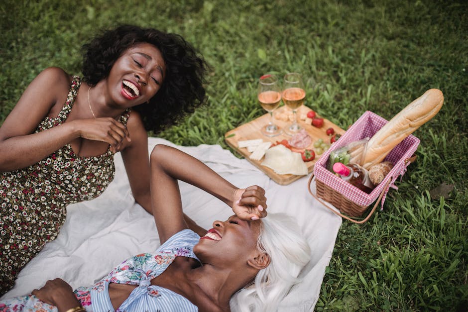 Two women enjoying a joyful picnic with wine and snacks on a lush green meadow.