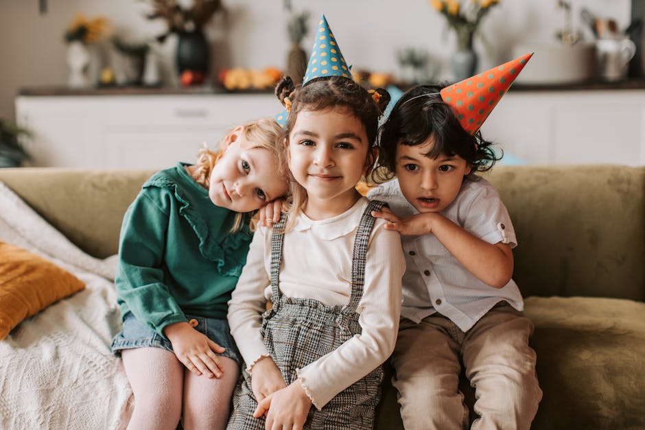 Three children in party hats enjoying a fun indoor celebration together on the couch.