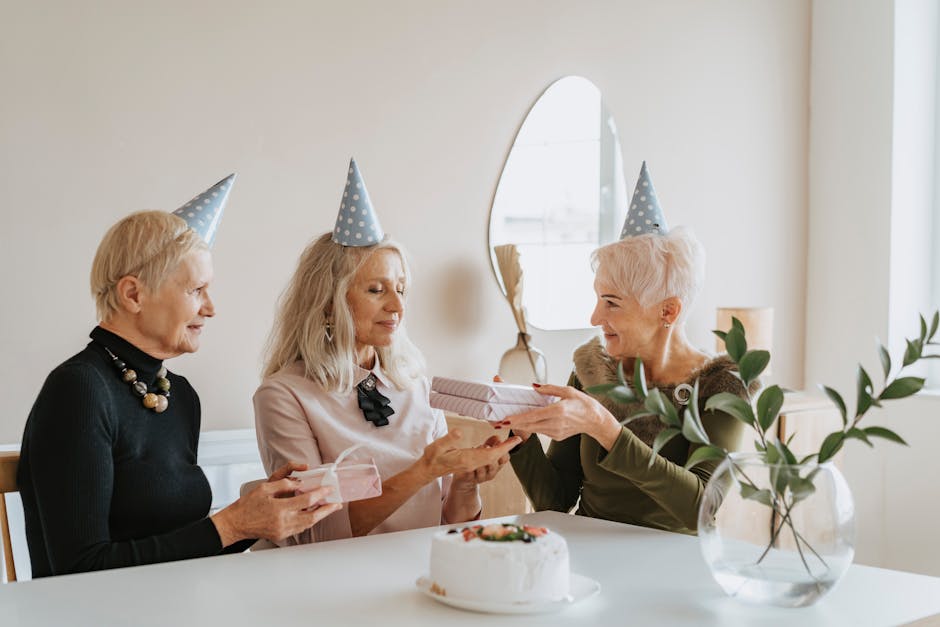 Three senior women celebrating a birthday with gifts, party hats, and cake indoors.