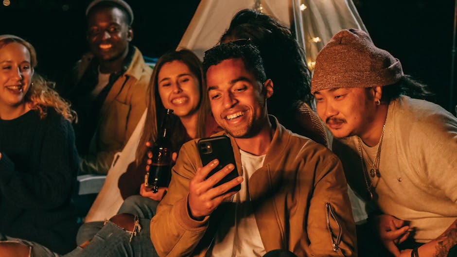 Group of friends smiling and enjoying a cozy campfire evening with a smartphone.