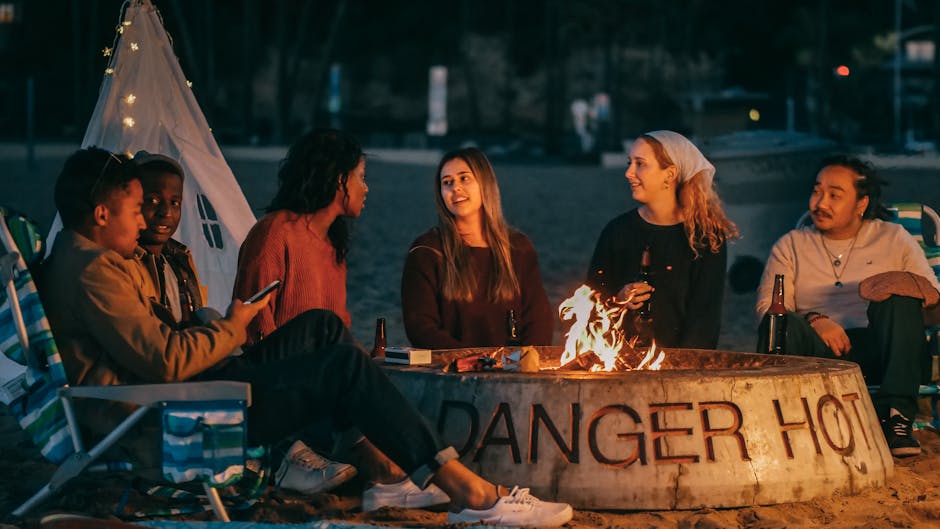 A diverse group of friends socializing around a beach bonfire at sunset, creating a warm atmosphere.