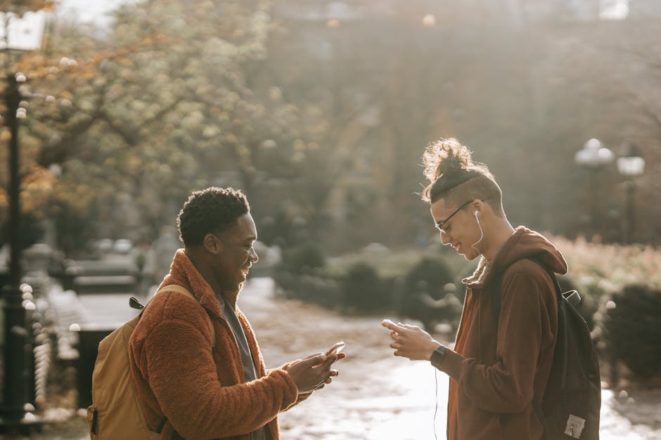 Side view of millennial laughing multiethnic friends wearing similar clothes using modern cellphones while standing in city park on autumn day