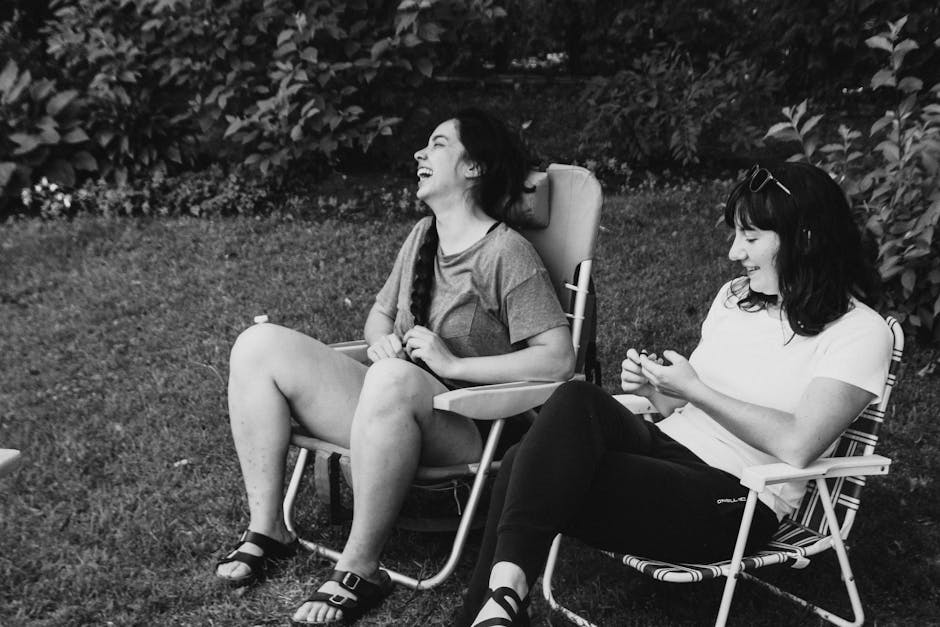 Two women enjoying a cheerful moment while sitting in folding chairs on a grass lawn.