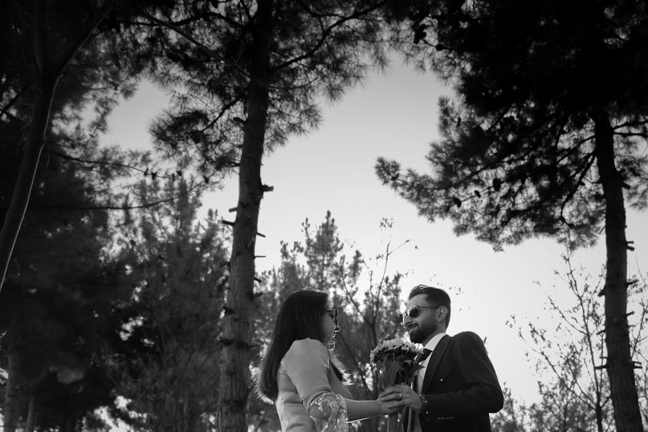 A romantic moment captured in black and white featuring a couple in a forest, holding a bouquet.