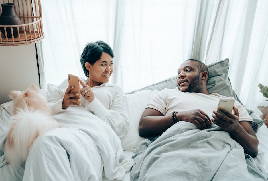 A couple bonding with their dog while using smartphones in a cozy bedroom setting.