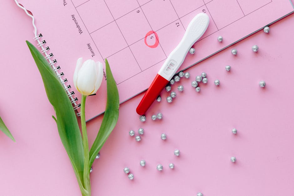 A close-up of a pregnancy test, calendar, and tulip arranged on a pink surface.