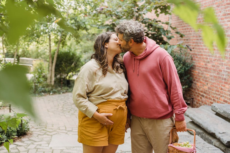 Happy pregnant woman in casual clothes touching tummy while kissing adult husband holding basket with fresh fruits during holidays in countryside