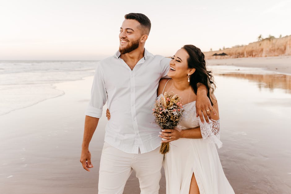 Smiling bearded groom in white shirt and trousers walking along sandy beach together with joyful bride in wedding dress