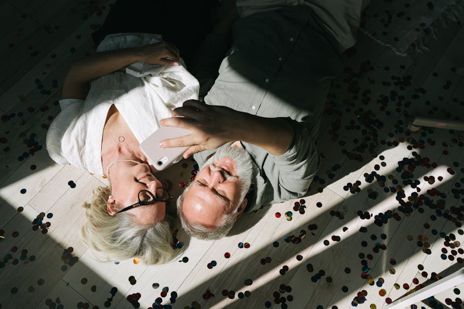 Happy senior couple lying on confetti-filled floor, taking a selfie and celebrating together.