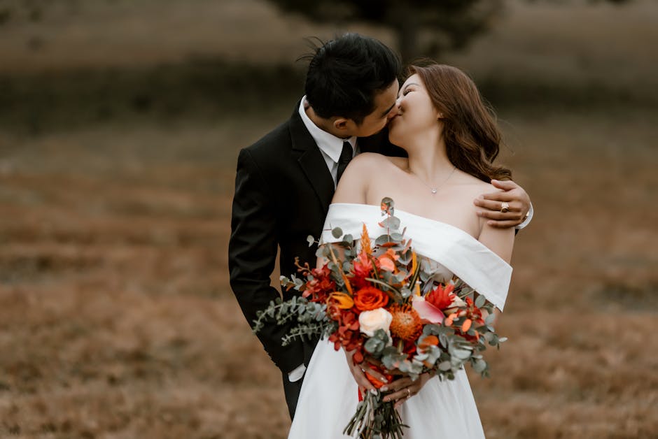 Loving Asian groom and bride with bouquet of flowers wearing wedding outfits kissing while standing in nature with blurred background in daylight
