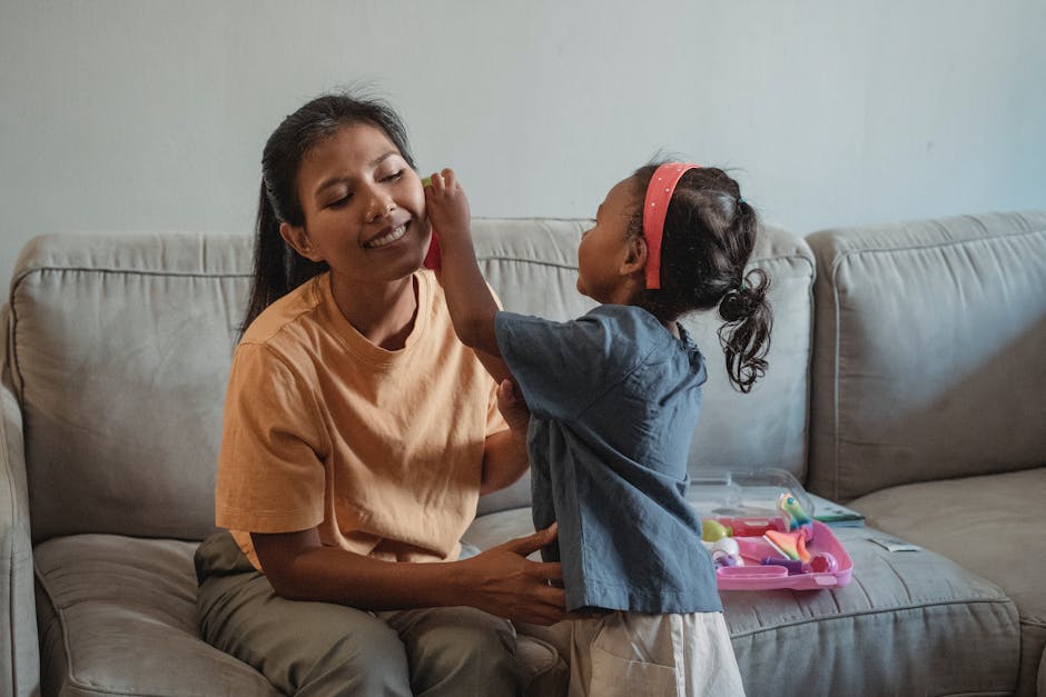 Playful little girl with toy near ear of cheerful mother doing check up while playing game on sofa in apartment