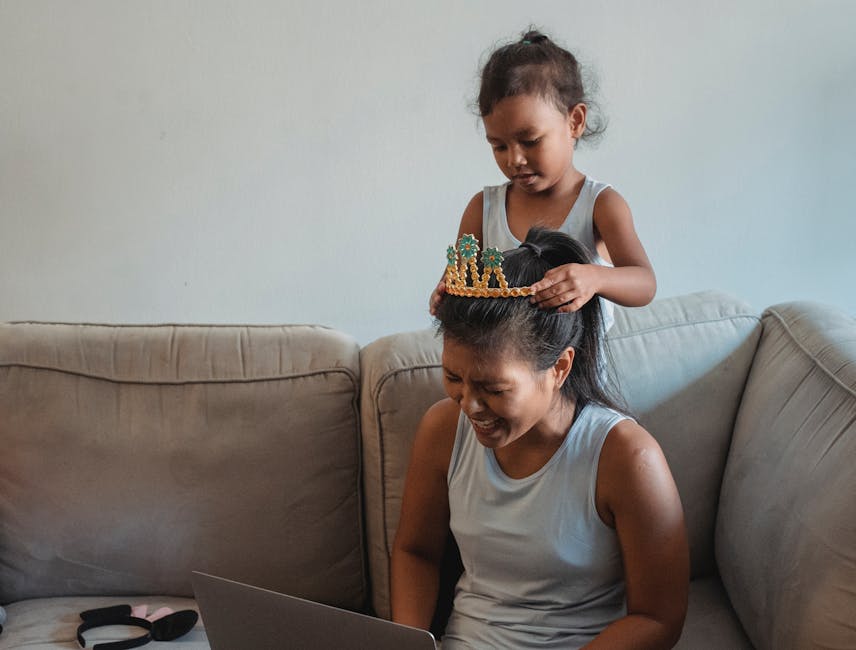 Adorable Asian girl putting crown on laughing mothers head siting on cozy couch and working on laptop at home