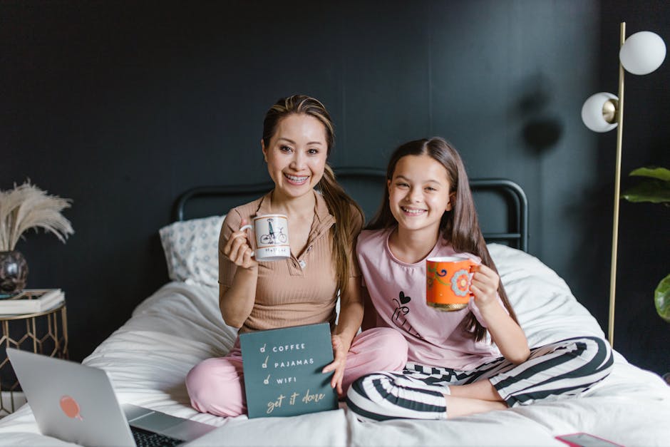A smiling mother and daughter relaxing on bed with coffee mugs and a tablet, portraying a warm family moment indoors.