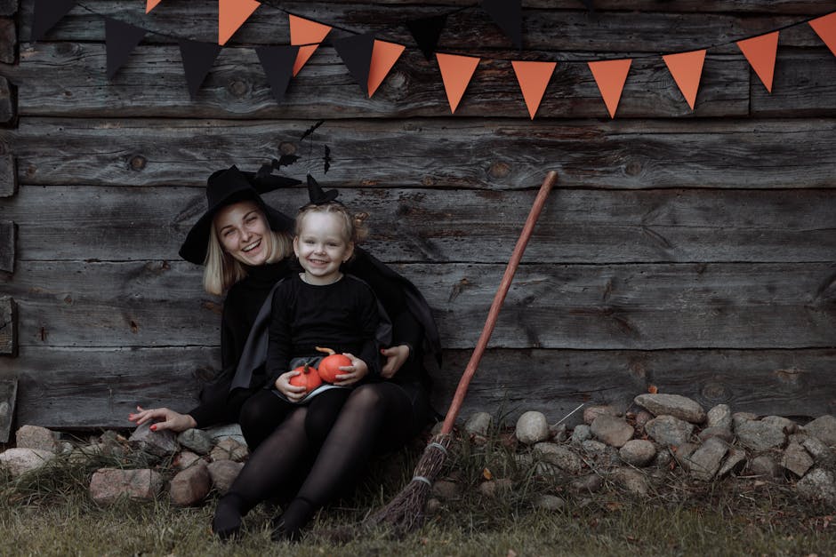 Smiling woman and child dressed as witches sitting outdoors with pumpkins and Halloween decorations.