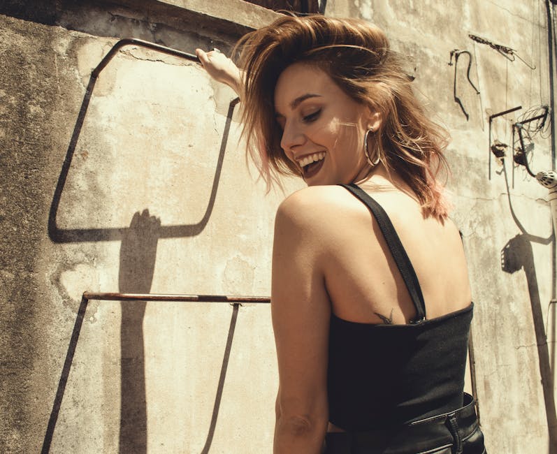 Young woman laughing and posing in a stylish outfit on a sunny day against an industrial backdrop.