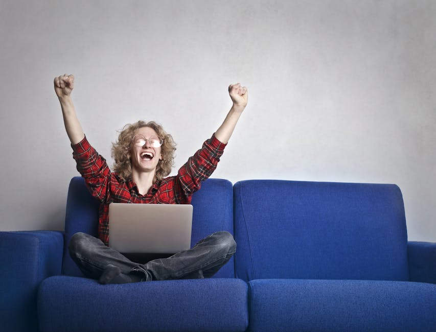 Thrilled individual celebrating success while using a laptop on a blue sofa indoors.