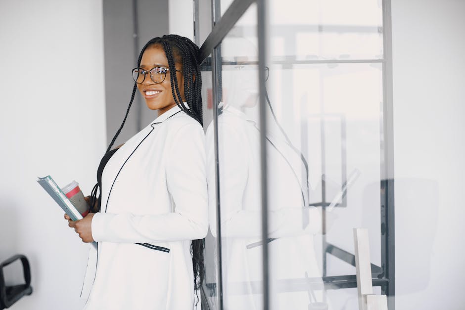 Smiling woman with braided hair leaning against glass panel holding books and coffee cup.