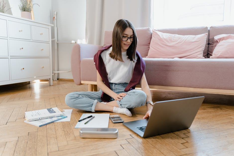 Teen girl studying online at home, sitting on the floor with a laptop and documents.