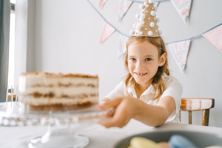 A joyful child wearing a party hat sits at a table with a birthday cake, ready to celebrate indoors.