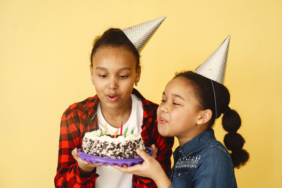 Two children in party hats joyfully blowing out birthday candles on a cake against a bright yellow background.