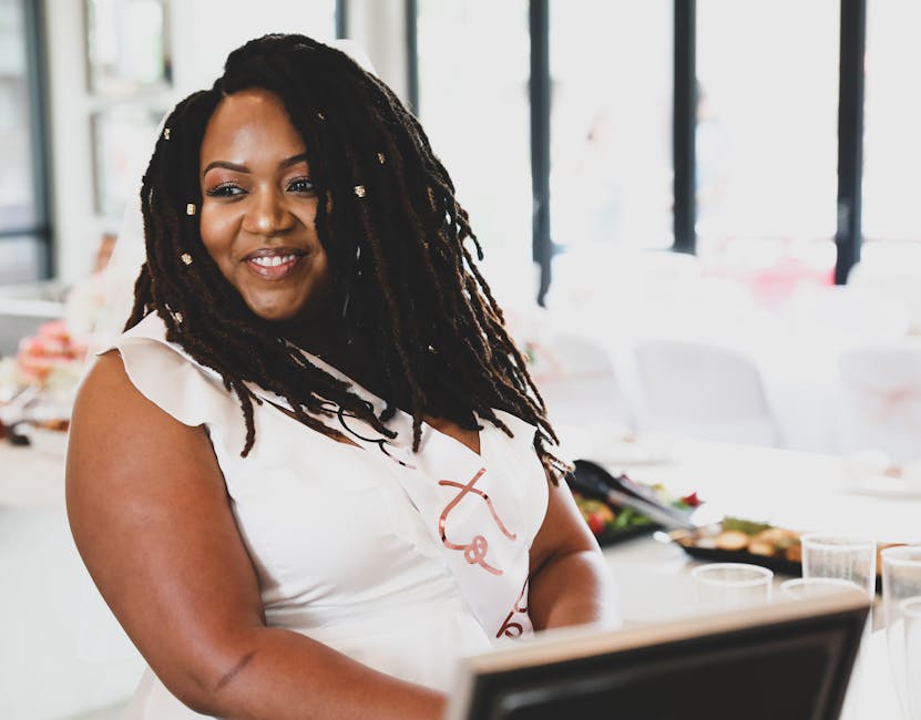 Black woman with dreadlocks smiling at an indoor celebration wearing a white dress with a sash.