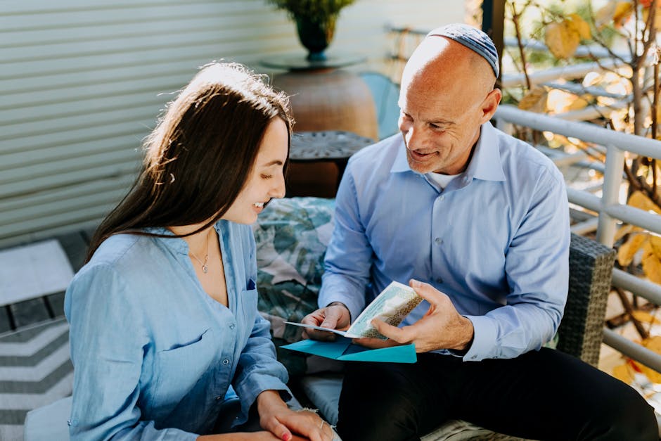 A father and daughter share a joyful Hanukkah moment with a card in a sunlit outdoor setting.