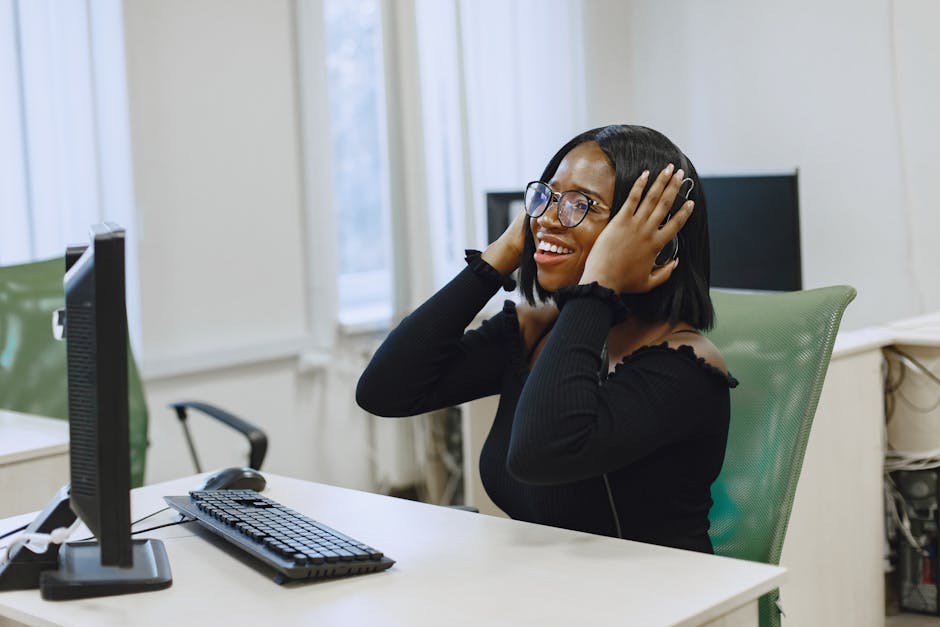 A cheerful woman enjoying music with a headset while sitting at a desk in an office setting.